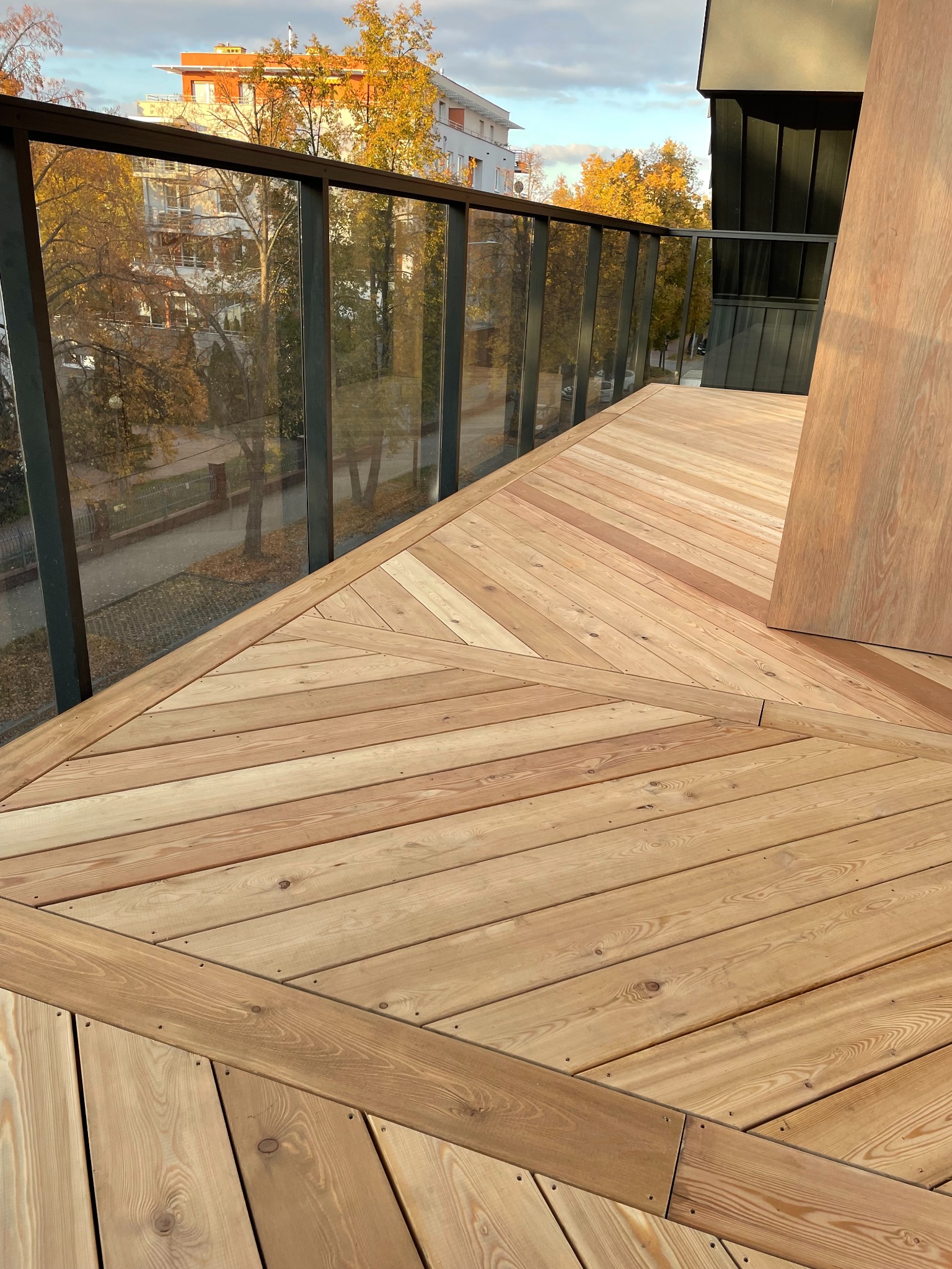 Nowoczesny balkon z drewnianą podłogą ułożoną we wzór jodełki, szklana balustrada z widokiem na jesienne drzewa. Naturalne oświetlenie podkreśla ciepły odcień drewna.