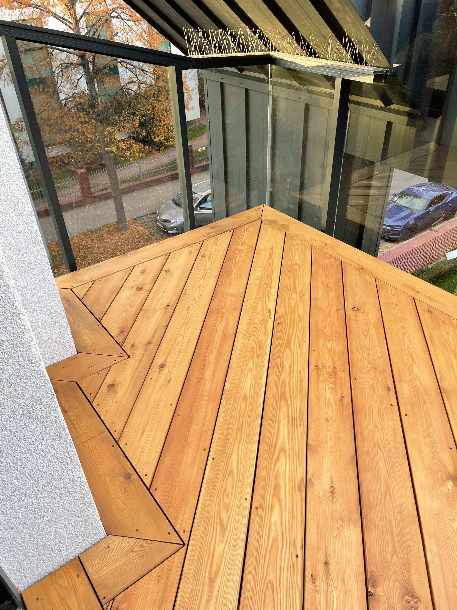 Wykończony taras z desek w odcieniach brązu, widok z góry, szklana balustrada z czarnymi elementami, jesienne drzewa w tle.