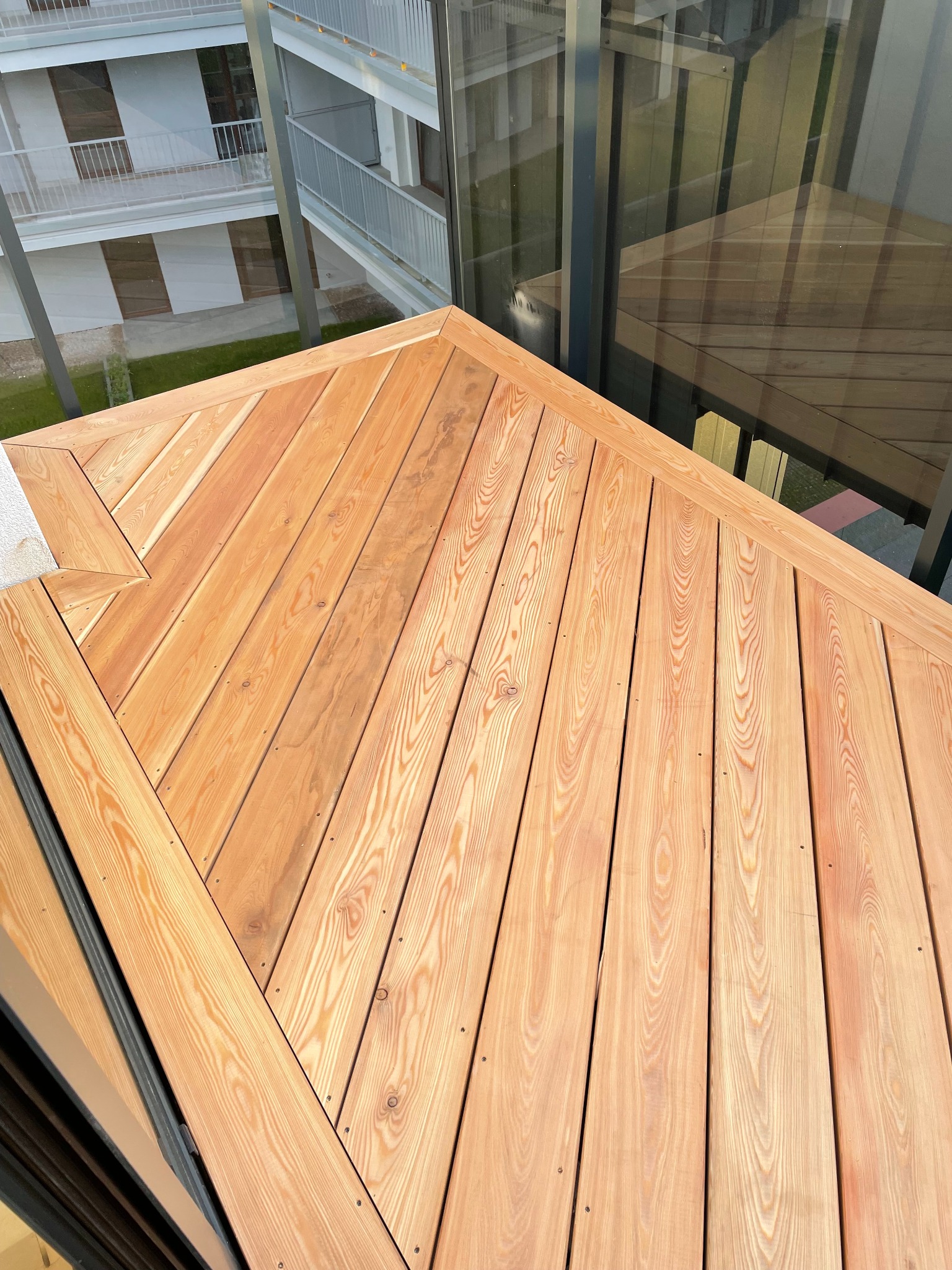 Ukończony, jasny taras z desek na balkonie apartamentu, widok z góry, z widocznym fragmentem elewacji i balustrady ze szkła. Deski ułożone ukośnie.