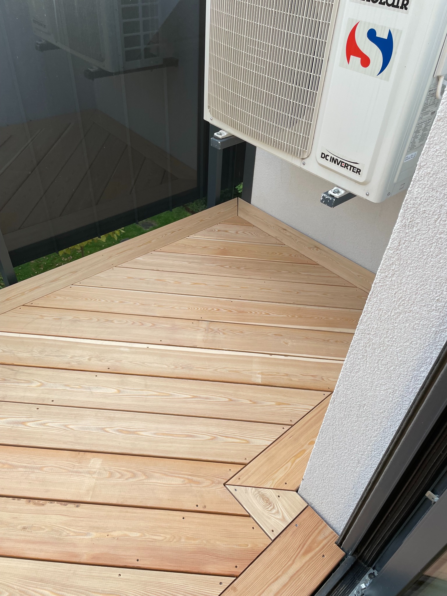 Jasny, drewniany taras w kształcie trójkąta na balkonie, z widocznym klimatyzatorem. Drewno ułożone równolegle, z delikatnym usłojeniem. Widok z góry.