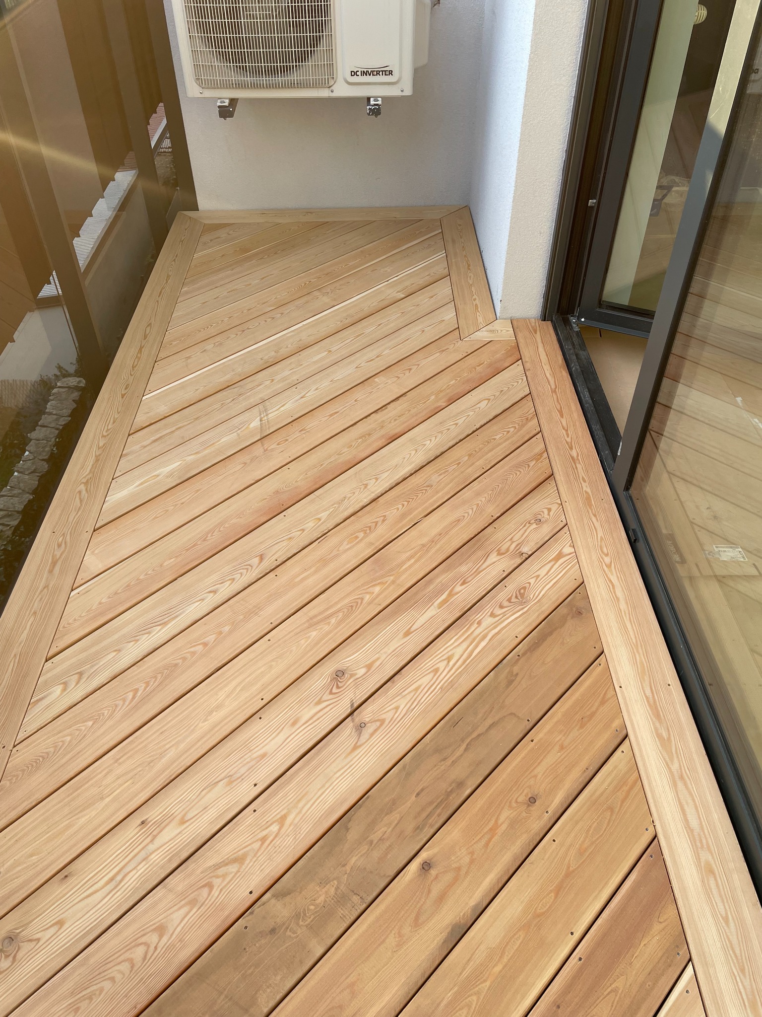 Jasny, ukośny wzór desek z naturalnego drewna tworzy podłogę tarasu przylegającego do budynku, z widocznym klimatyzatorem na ścianie. Czyste linie i jasne drewno.