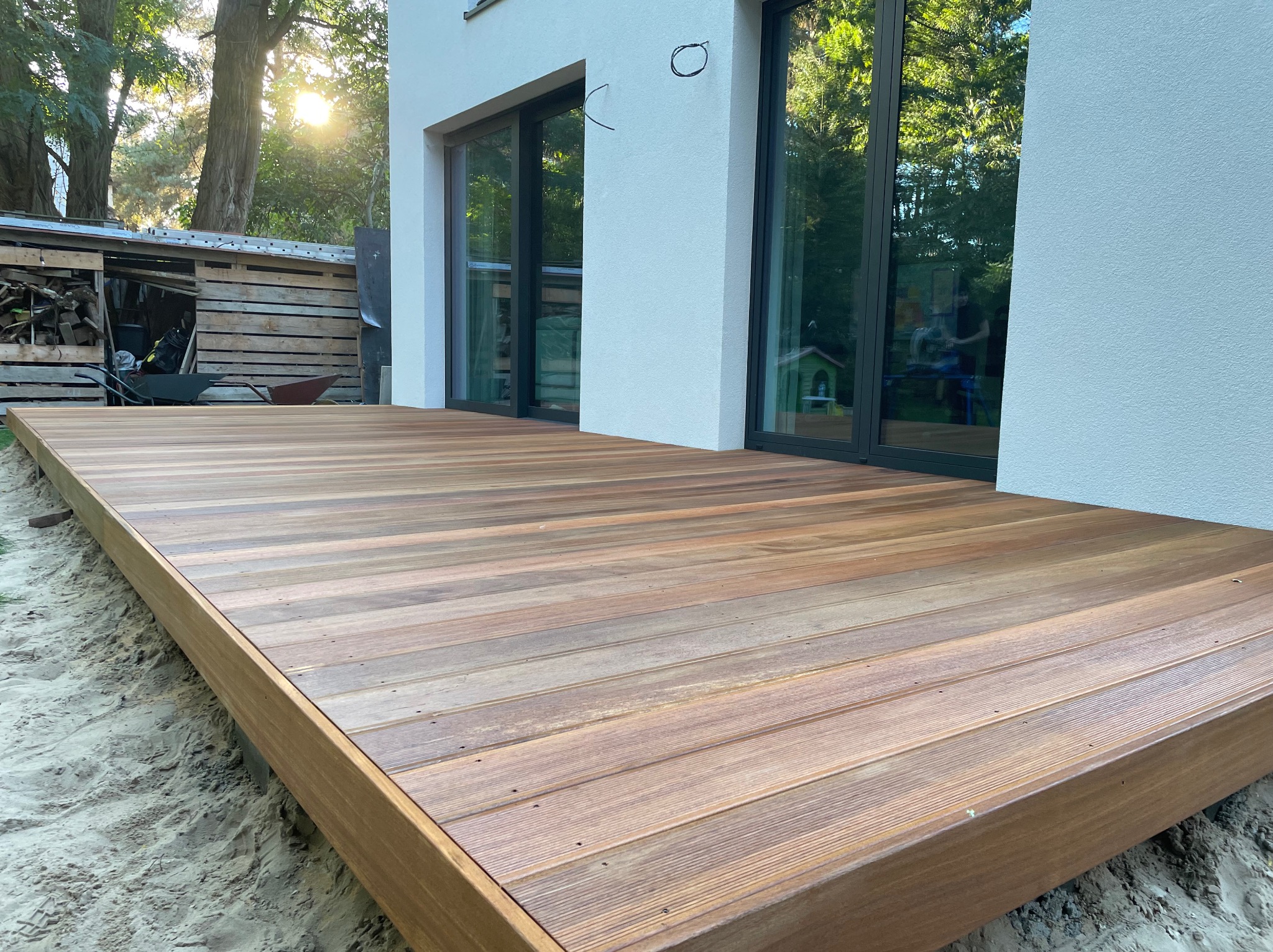 Nowoczesny taras z desek o ciepłym odcieniu brązu, przylegający do białego budynku z dużymi oknami. Piasek wokół tarasu, zieleń w tle.