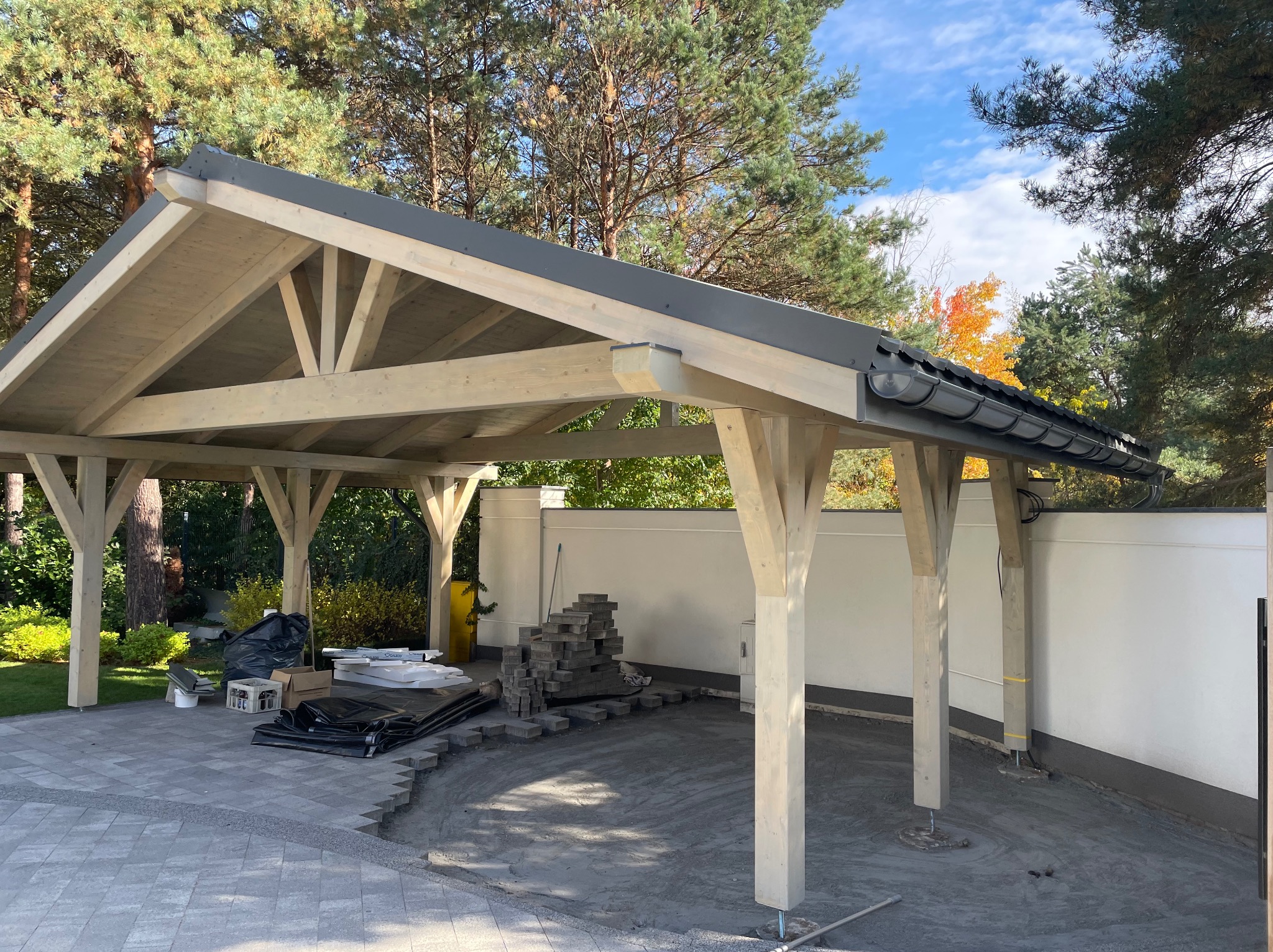 Drewniana konstrukcja z szarym dachem w ogrodzie, w trakcie budowy. Widoczne materiały budowlane i częściowo ułożona kostka brukowa.