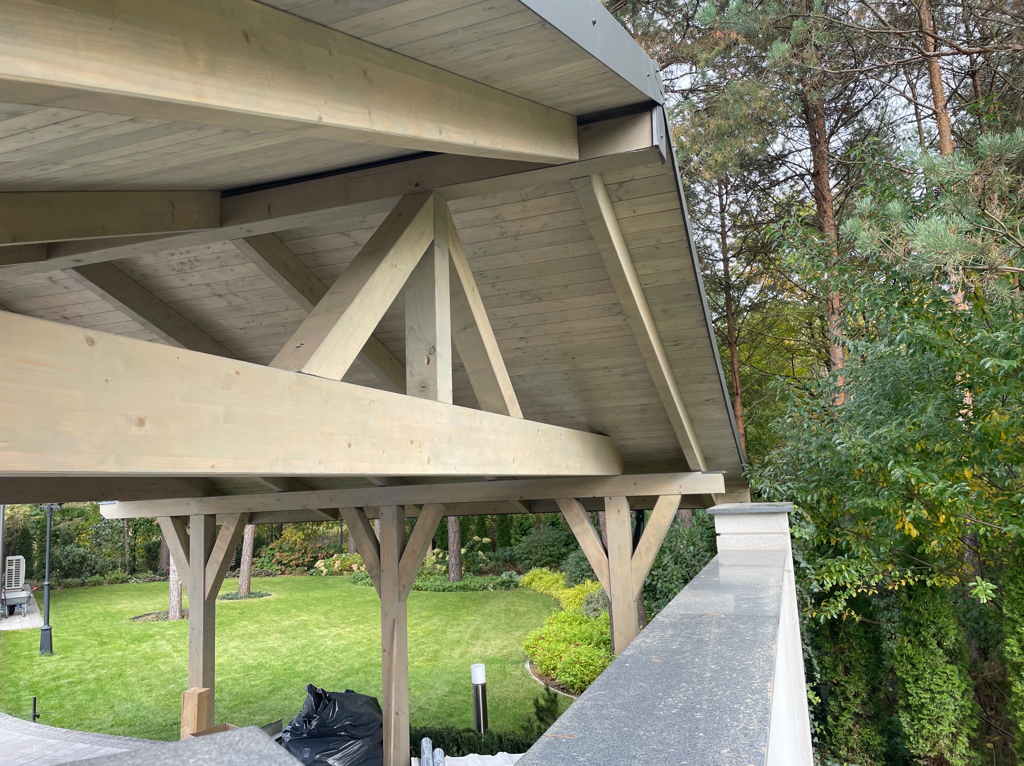 Drewniana konstrukcja zadaszenia ogrodu z widoczną więźbą dachową i słupami nośnymi, otoczona zielenią. Elementy konstrukcyjne pomalowane na jasny kolor.