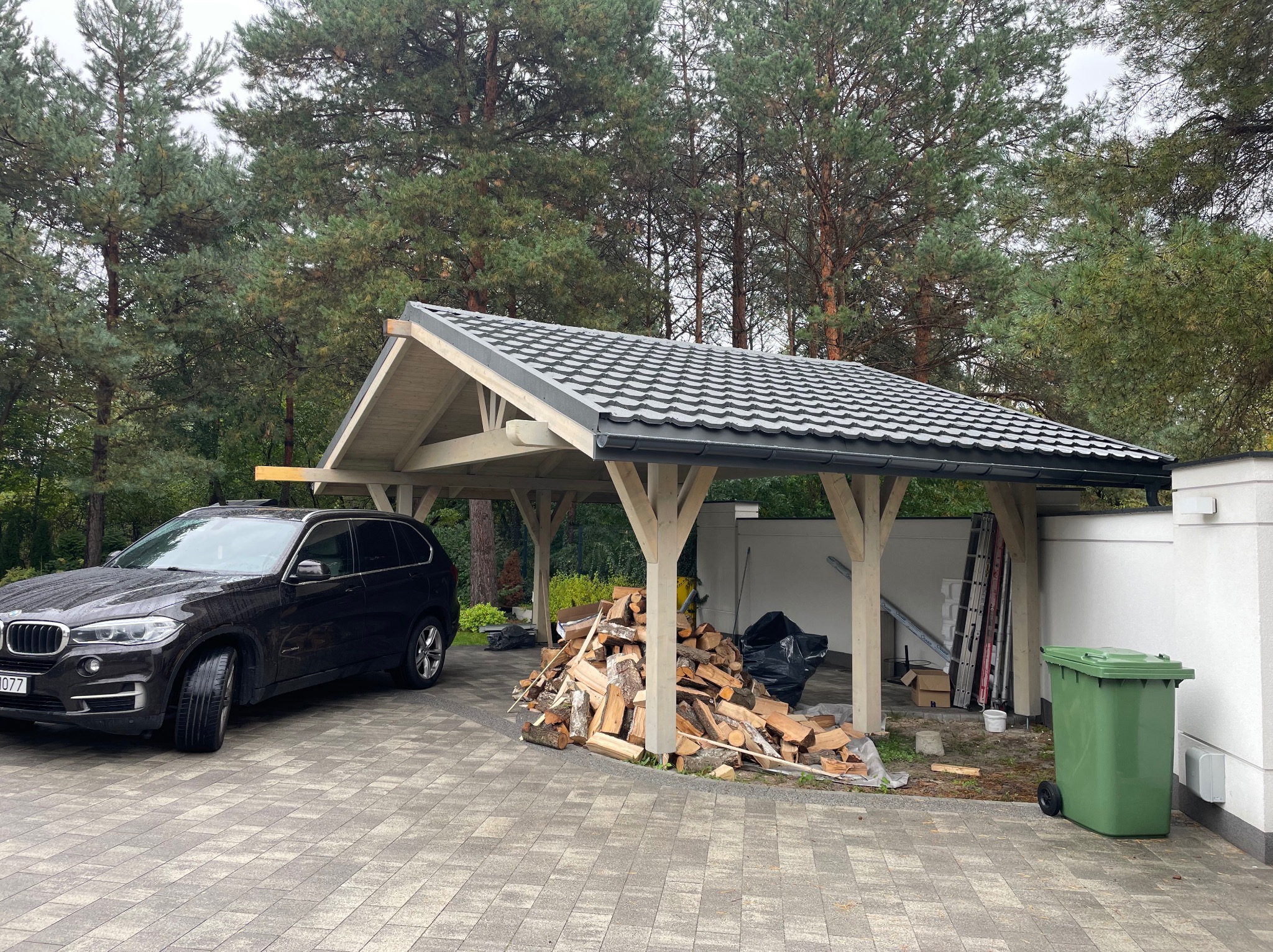 Nowoczesna, drewniana wiata garażowa z szarą dachówką i stosem drewna opałowego obok, na tle drzew i kostki brukowej.