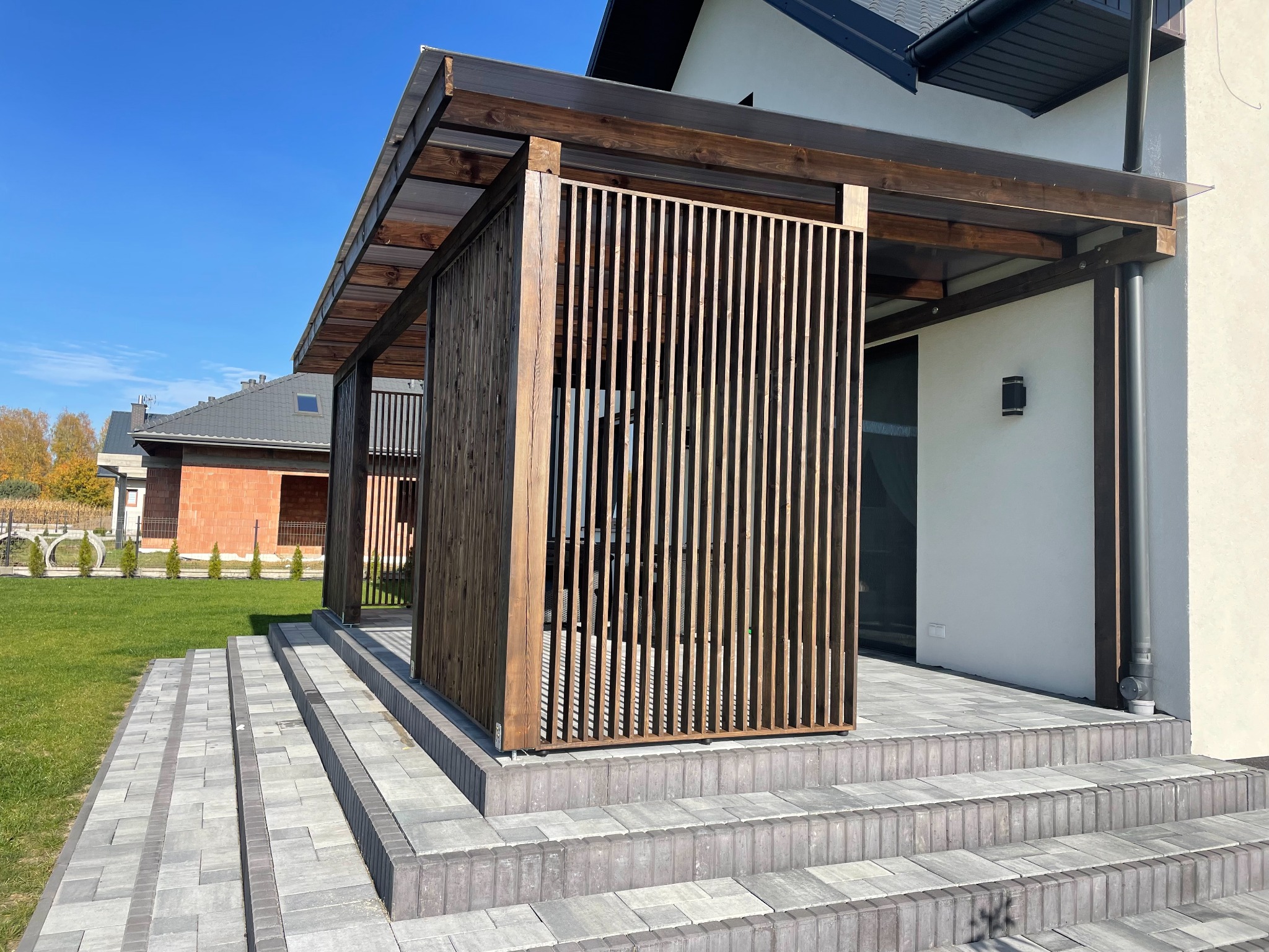 Drewniana pergola z ażurowymi ściankami, zadaszona przezroczystym materiałem, na tarasie z szarej kostki brukowej. Nowoczesny design, błękitne niebo w tle.