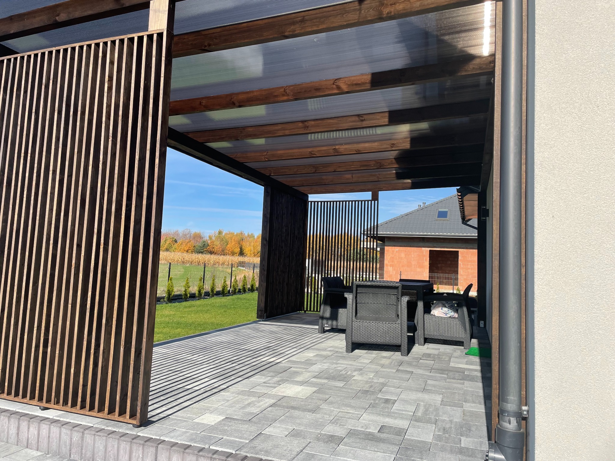 Zadaszona przestrzeń wypoczynkowa z drewnianą konstrukcją i ażurowymi ściankami, wyposażona w meble ogrodowe na wyłożonym kostką tarasie.