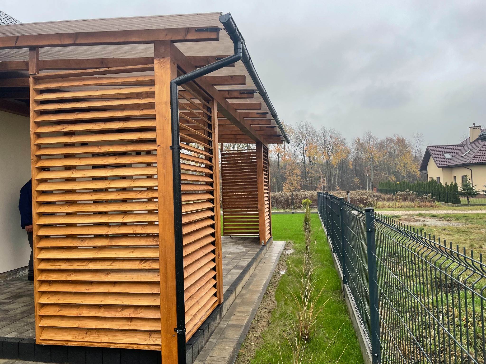 Nowoczesna, drewniana konstrukcja z lamelami i czarną rynną, widoczna z zewnątrz, na tle zieleni i domu. Przejrzysty dach. Ujęcie w pochmurny dzień.