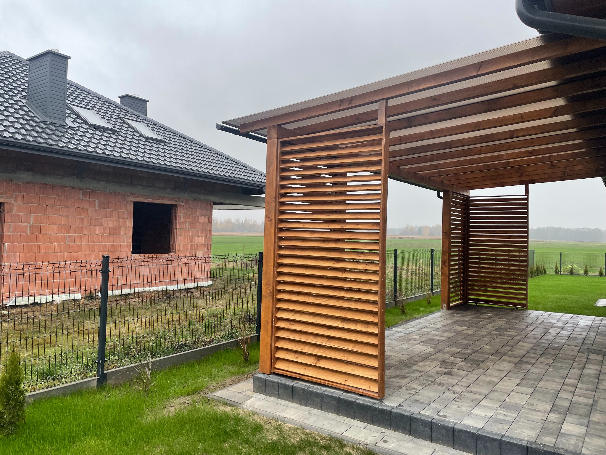 Nowoczesna, drewniana pergola z lamelami, przylegająca do domu w trakcie budowy. Uwagę przyciąga geometryczny wzór ułożonej kostki brukowej.