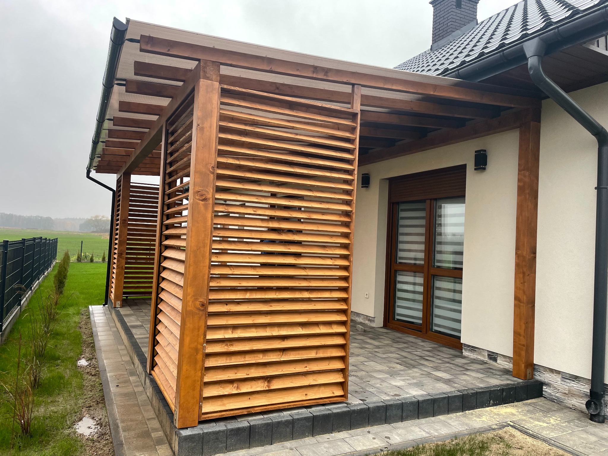 Drewniana pergola z lamelami w Warszawie, przylegająca do budynku z oknem i roletami. Zadaszenie z przezroczystych płyt. Kostka brukowa na podłożu.