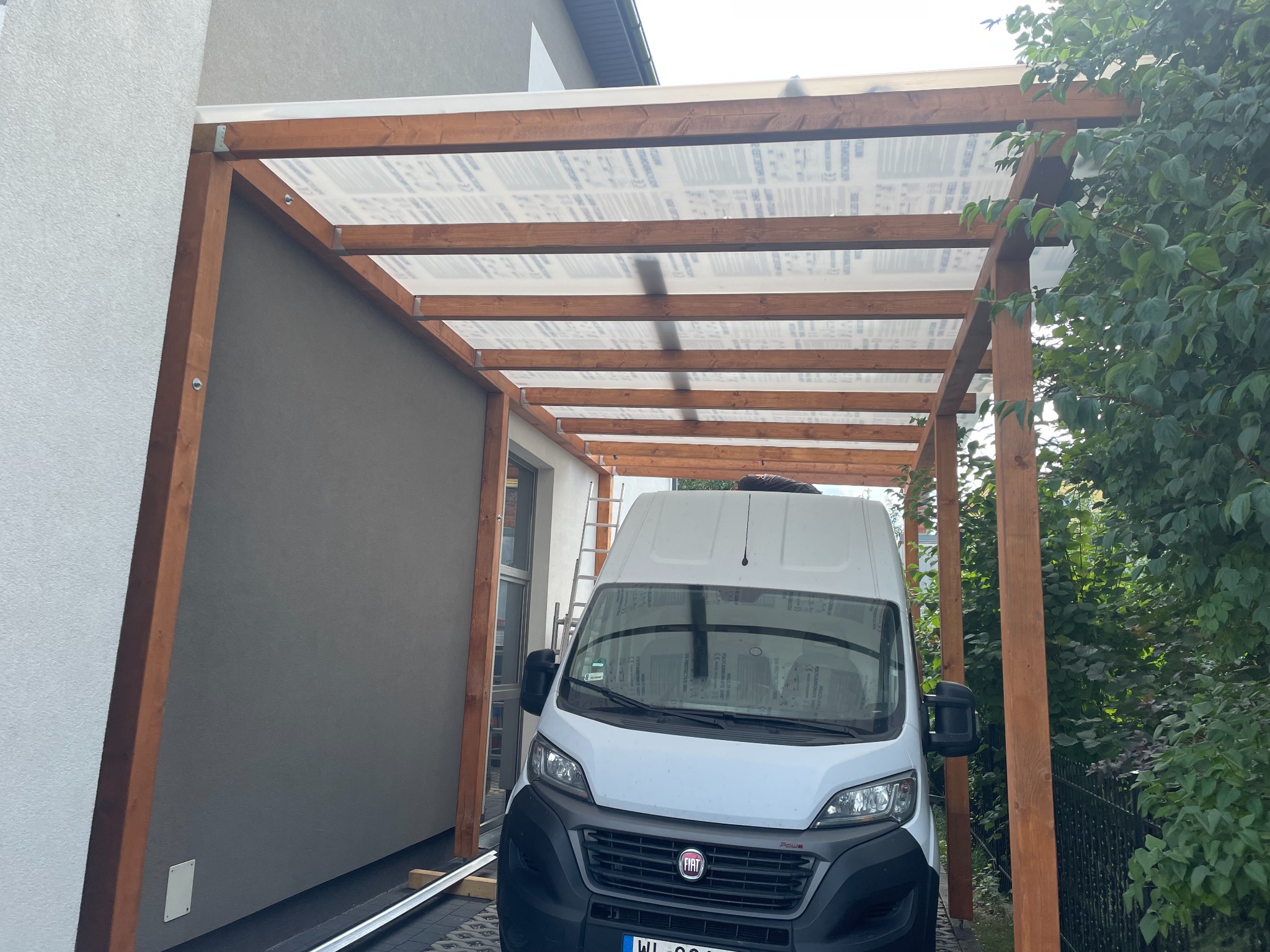 Drewniana wiata garażowa z przezroczystym zadaszeniem, częściowo przylegająca do budynku. Biały samochód dostawczy zaparkowany pod wiatą. Widoczne elementy konstrukcyjne i otoczenie.