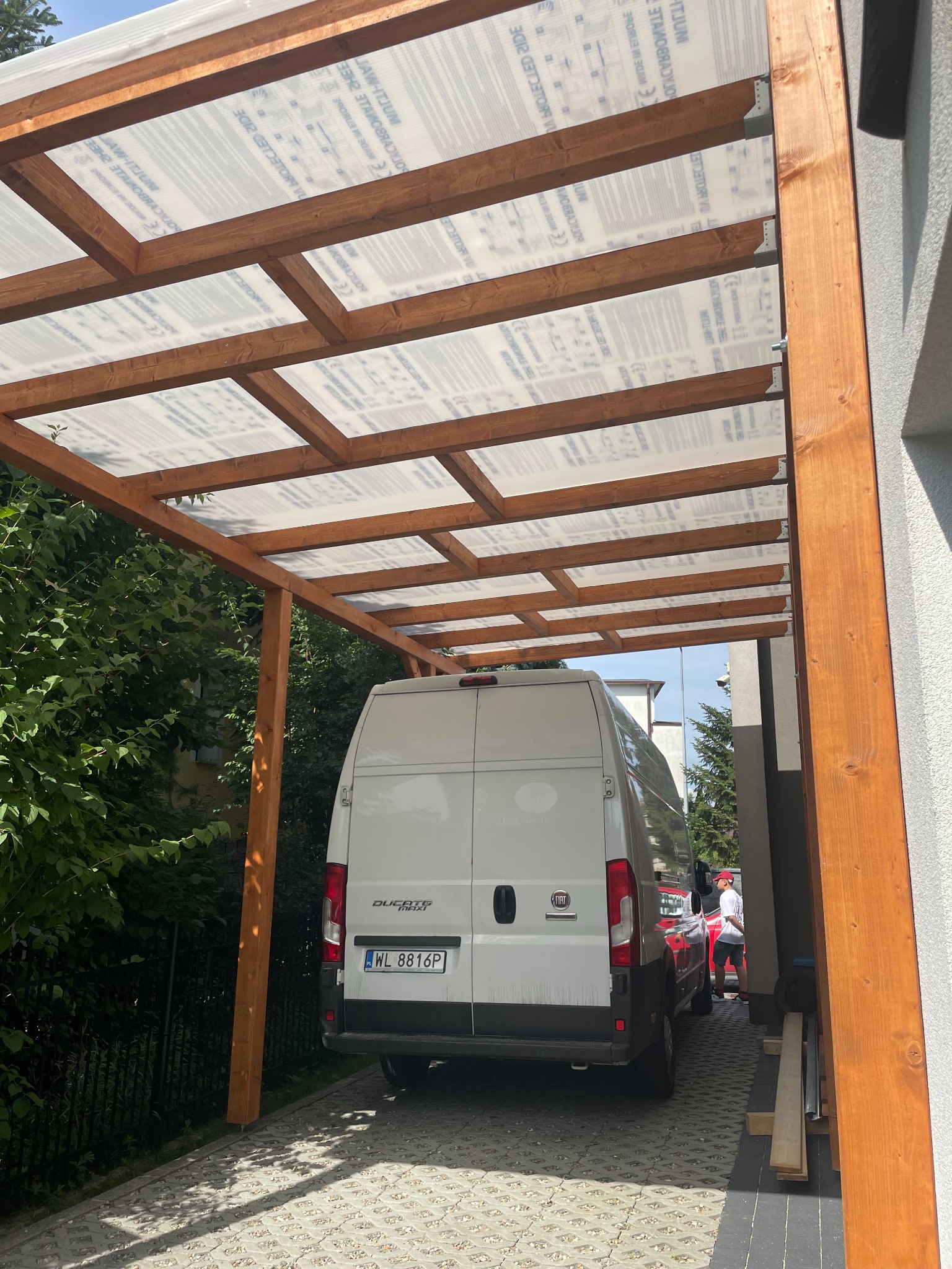 Drewniana konstrukcja z przezroczystym zadaszeniem, prawdopodobnie wiata garażowa, z zaparkowanym białym busem. Widok od dołu, z perspektywy parkingu.