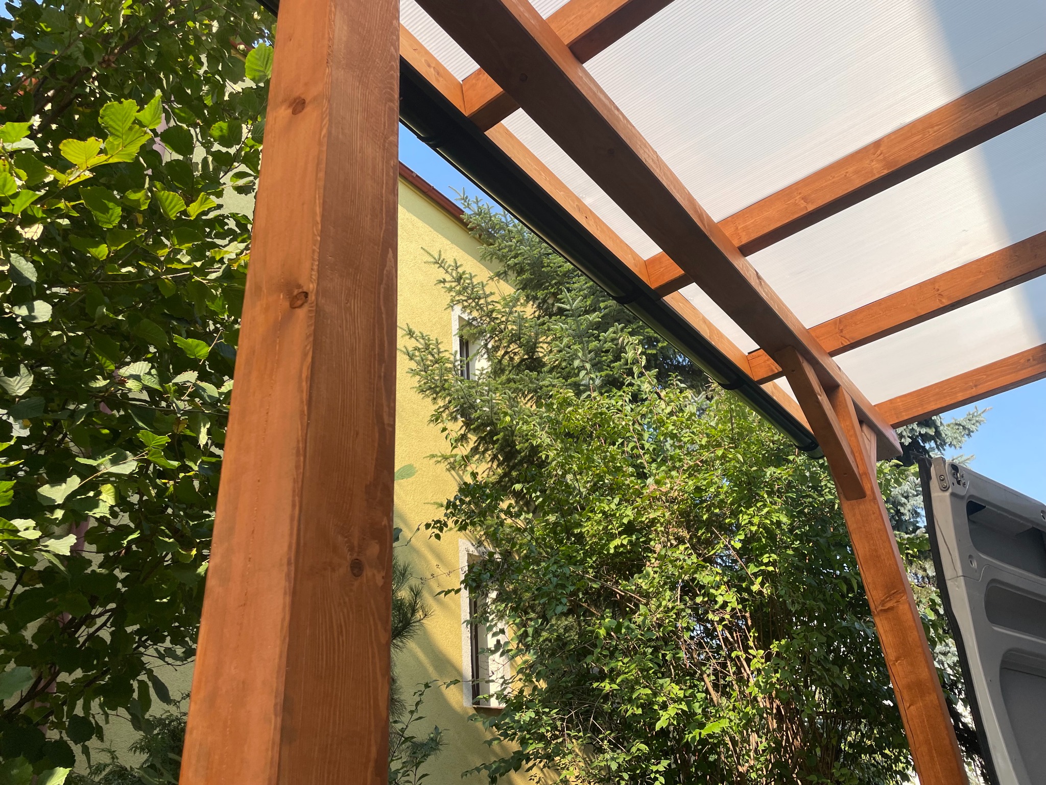 Drewniana konstrukcja z przezroczystym zadaszeniem, widok z dołu, zieleń w tle. Elementy konstrukcyjne z wyraźnym rysunkiem drewna. Jasne, naturalne oświetlenie.