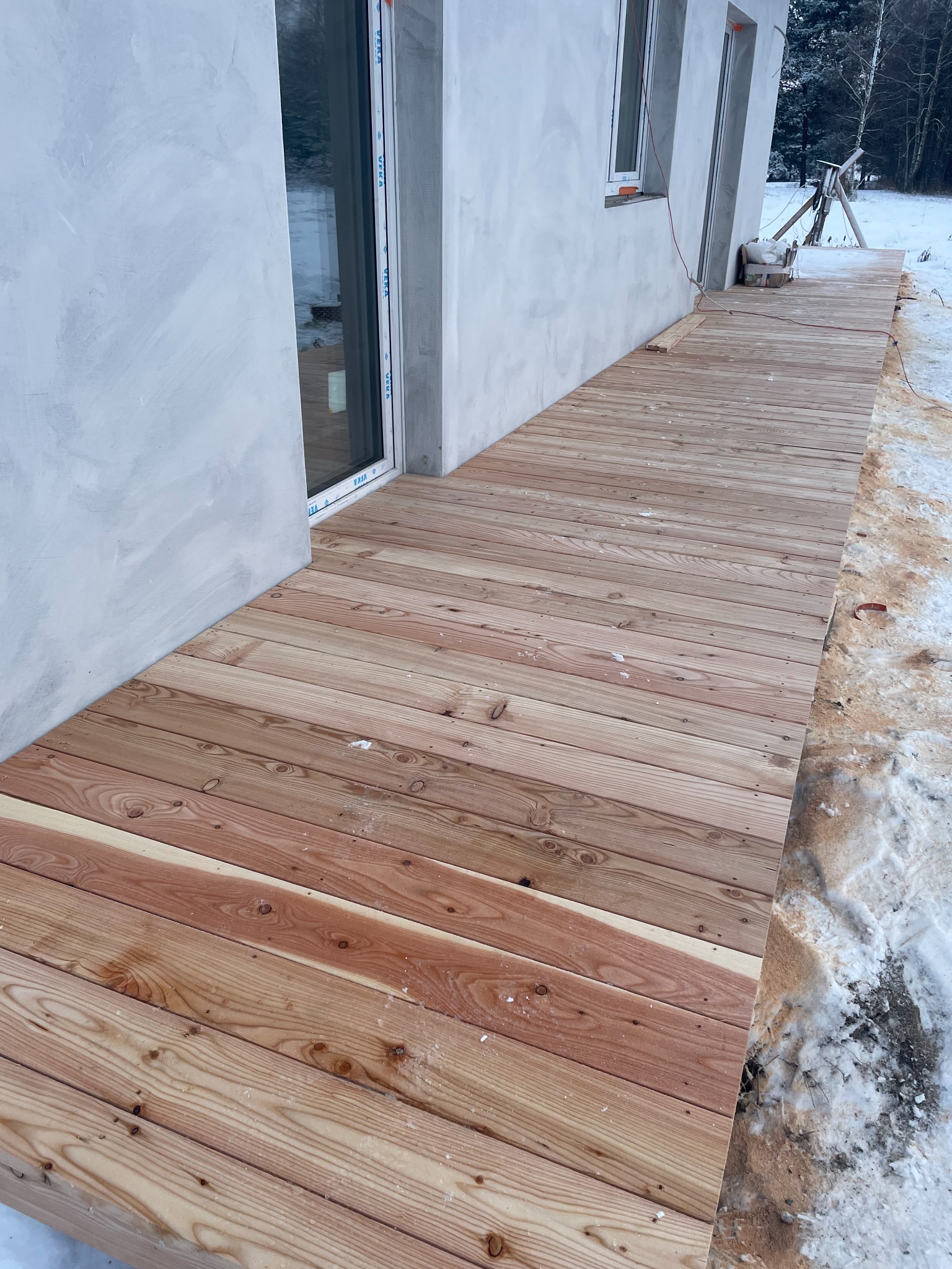 Nowy, jasny taras z desek przy budynku w zimowej scenerii. Widoczne świeże drewno i śnieg. Ujęcie z niskiej perspektywy podkreśla długość tarasu.
