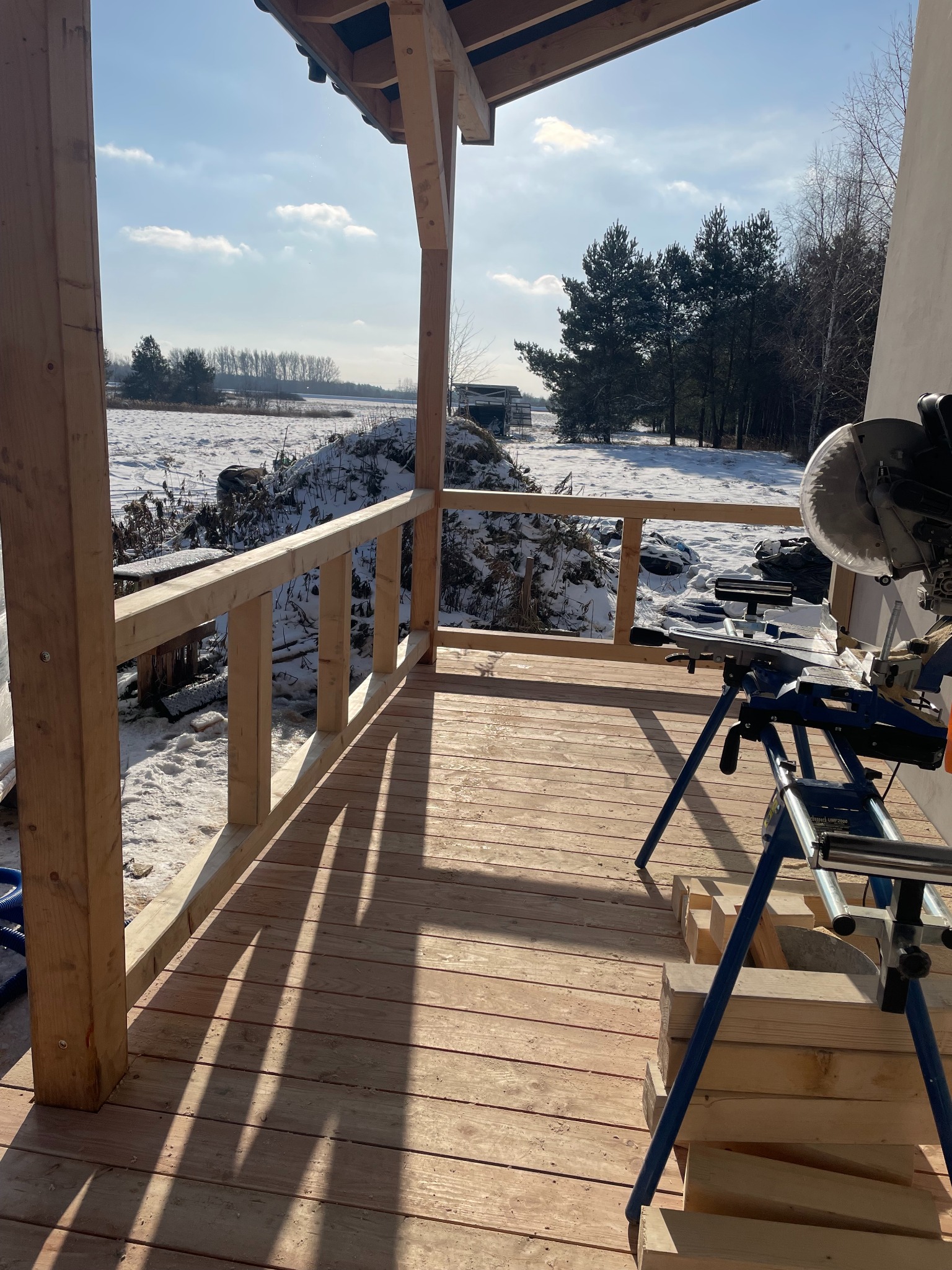 W trakcie budowy drewniany taras z widokiem na zimowy krajobraz. Widoczna piła do drewna i stos desek. Jasne drewno, słoneczny dzień.