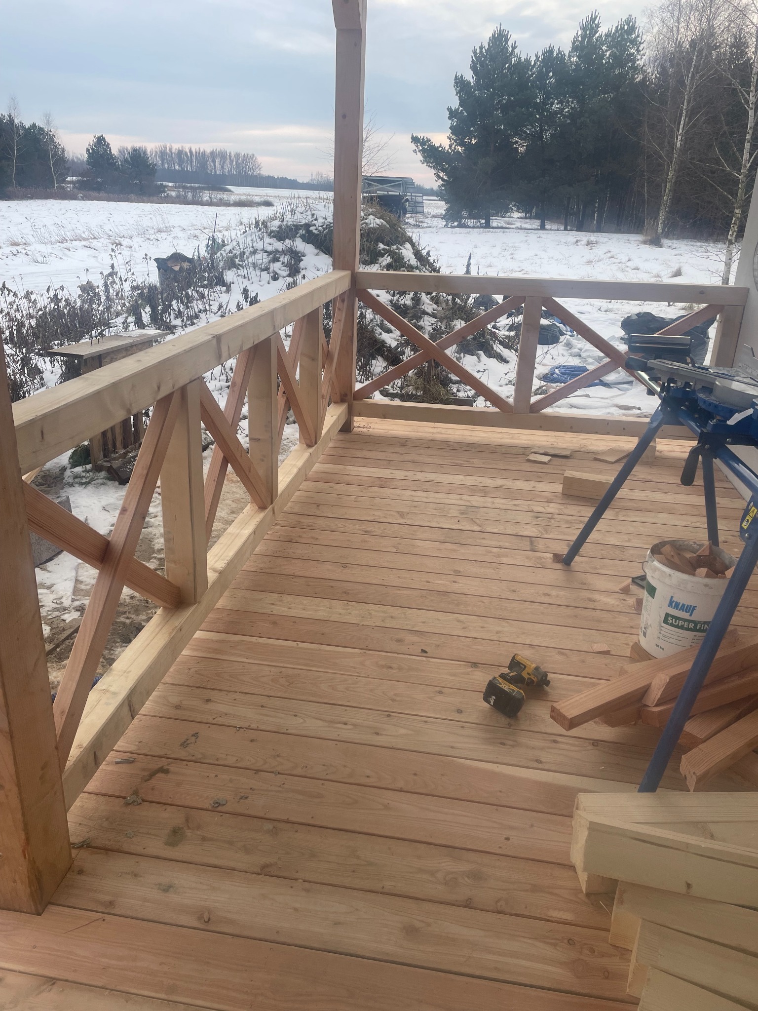 Drewniany taras w trakcie budowy w Warszawie, z widoczną konstrukcją balustrady i deskami podłogowymi. W tle zimowy krajobraz.