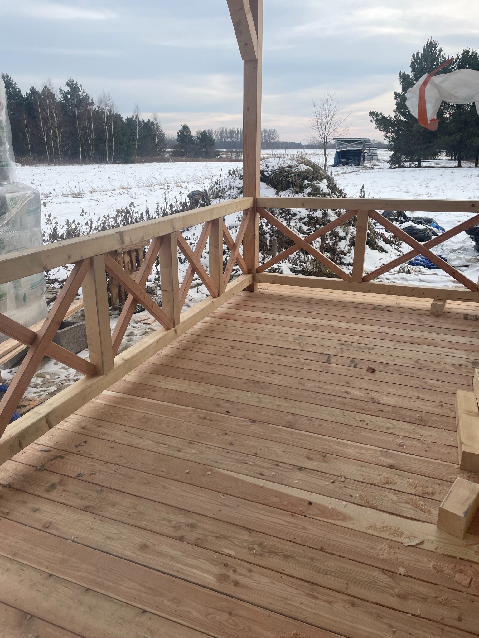 W zimowej scenerii, drewniany taras z balustradą w trakcie budowy. Widoczne deski podłogowe i elementy konstrukcyjne, a w tle ośnieżone pole i las.