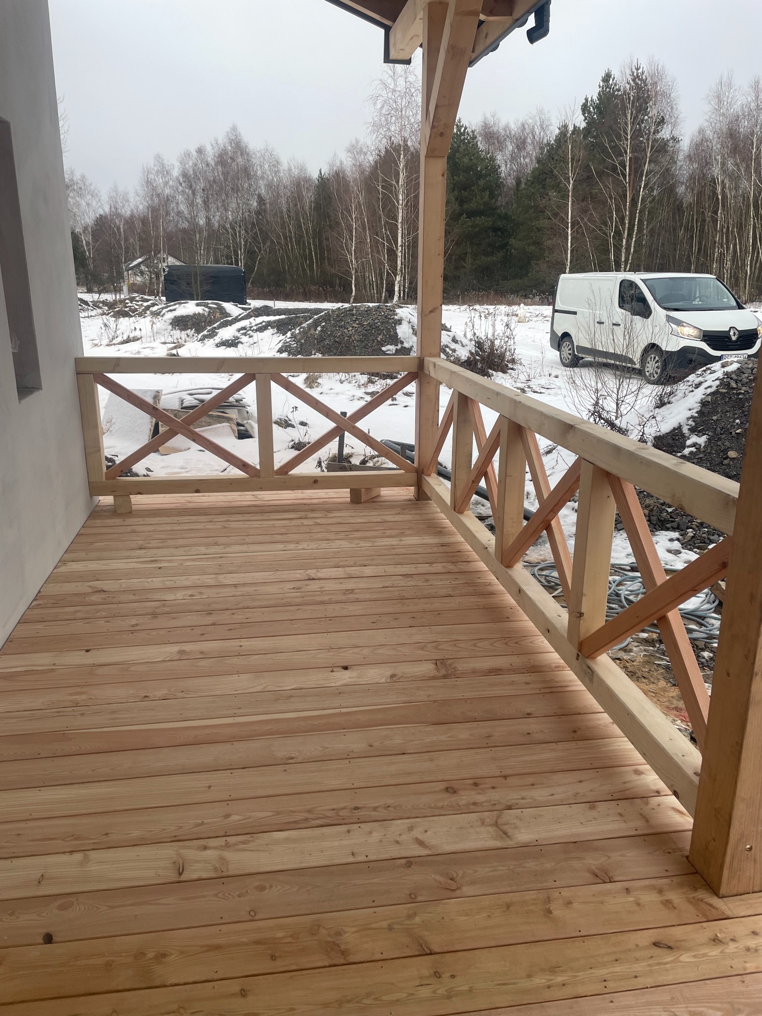 Nowy taras z jasnego drewna z balustradą w kształcie krzyży, na tle zimowego krajobrazu i białego samochodu dostawczego.