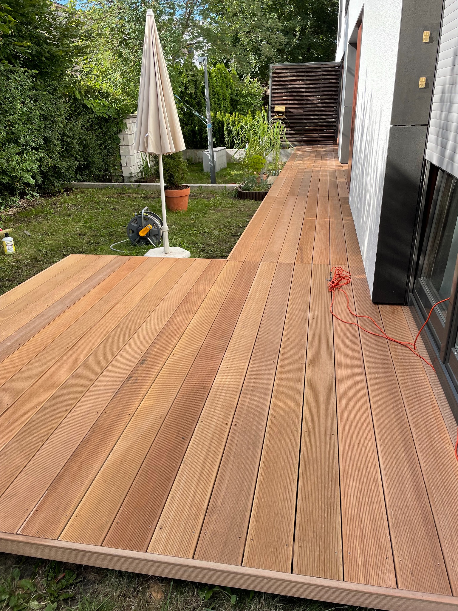 Nowoczesny taras z desek kompozytowych w odcieniach brązu, przylegający do budynku z jasną elewacją. Widoczny parasol ogrodowy i zadbana zieleń w tle.