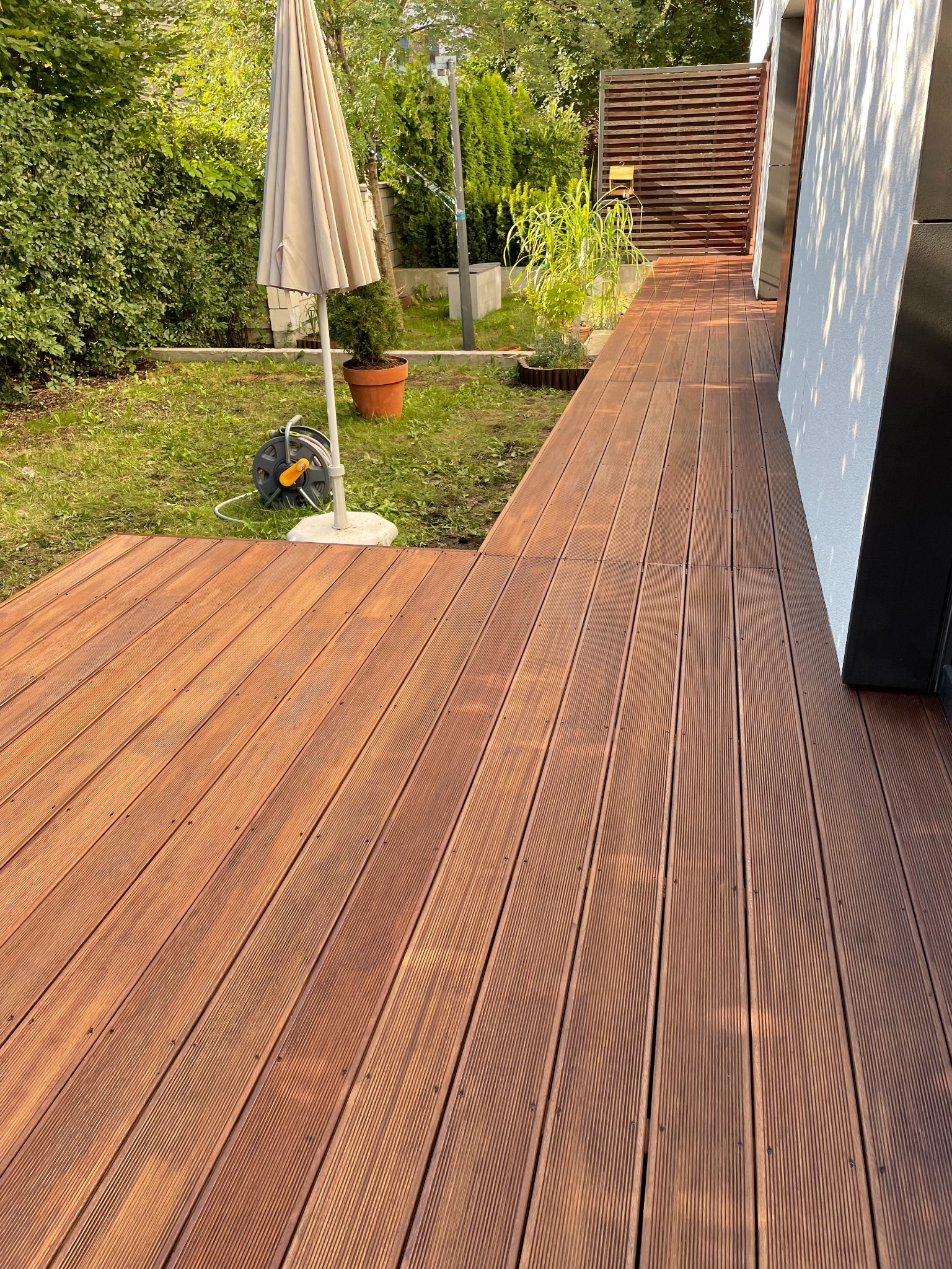 Nowoczesny taras z desek kompozytowych w ogrodzie, z widocznym parasolem i roślinami w donicach. Czyste linie, ciepłe oświetlenie, zadbana przestrzeń.