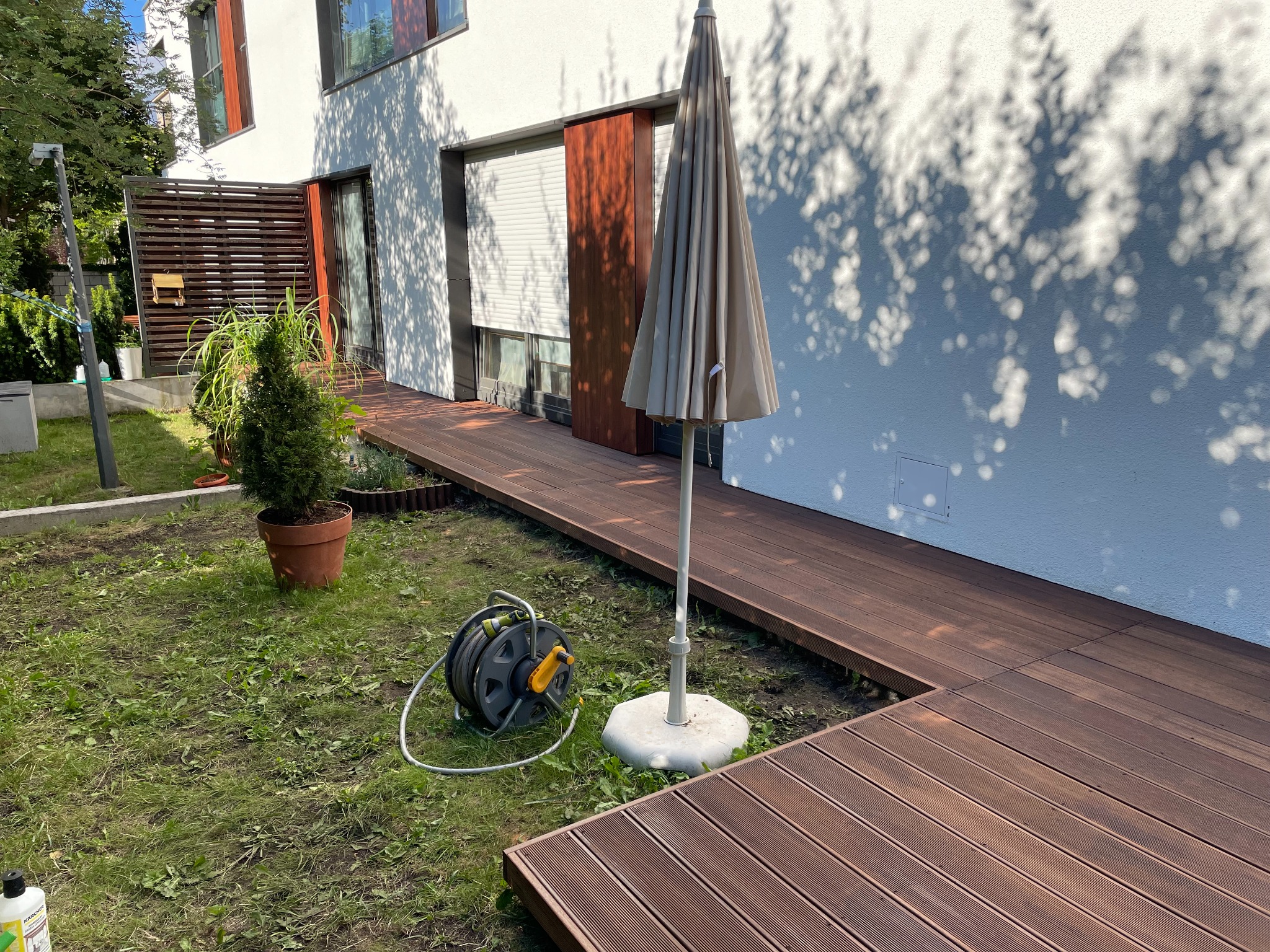 Nowoczesny taras z desek kompozytowych w ogrodzie, z widocznym zwiniętym wężem ogrodowym i parasolem. Cień drzew na białej ścianie domu.