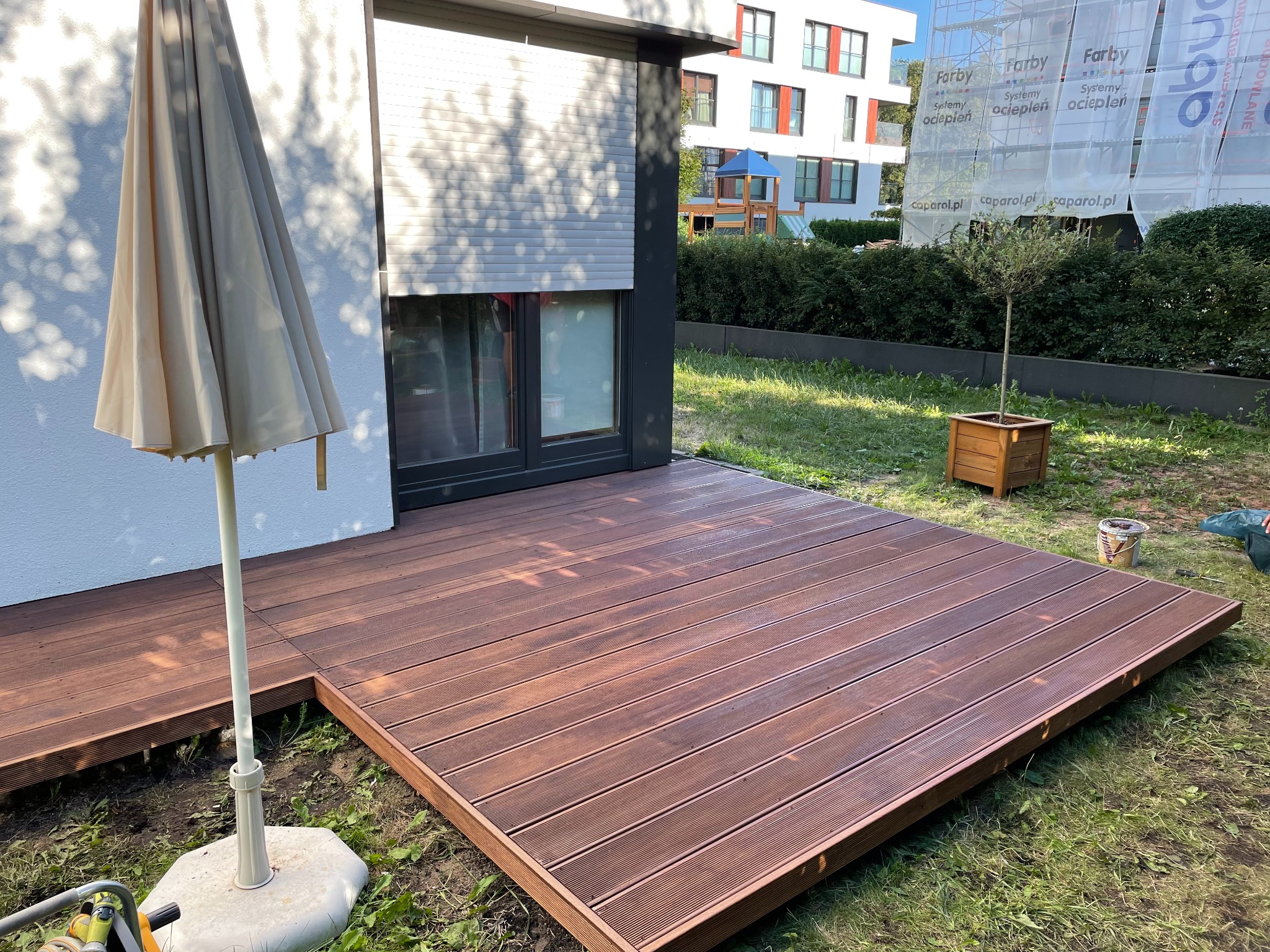 Nowoczesny taras z brązowych desek kompozytowych przylegający do budynku z roletami, z parasolem i widokiem na budynek w rusztowaniach.