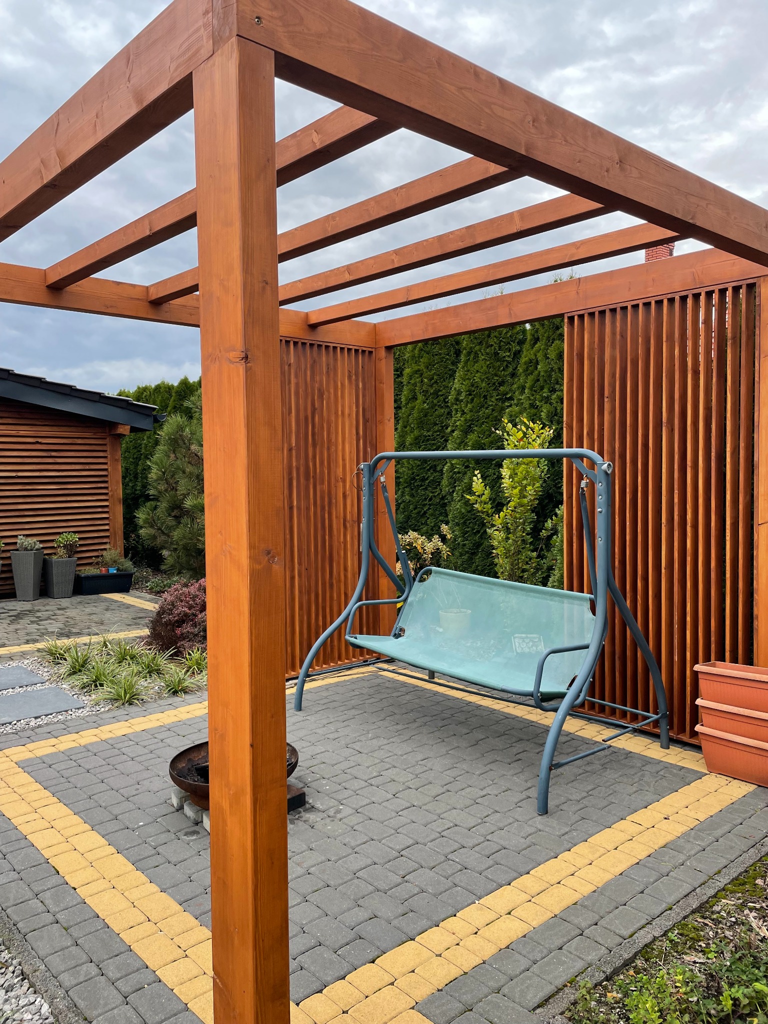 Drewniana pergola z huśtawką ogrodową na kostce brukowej w Warszawie. Konstrukcja z pionowymi lamelami, w tle zieleń i budynek z drewnianą elewacją.