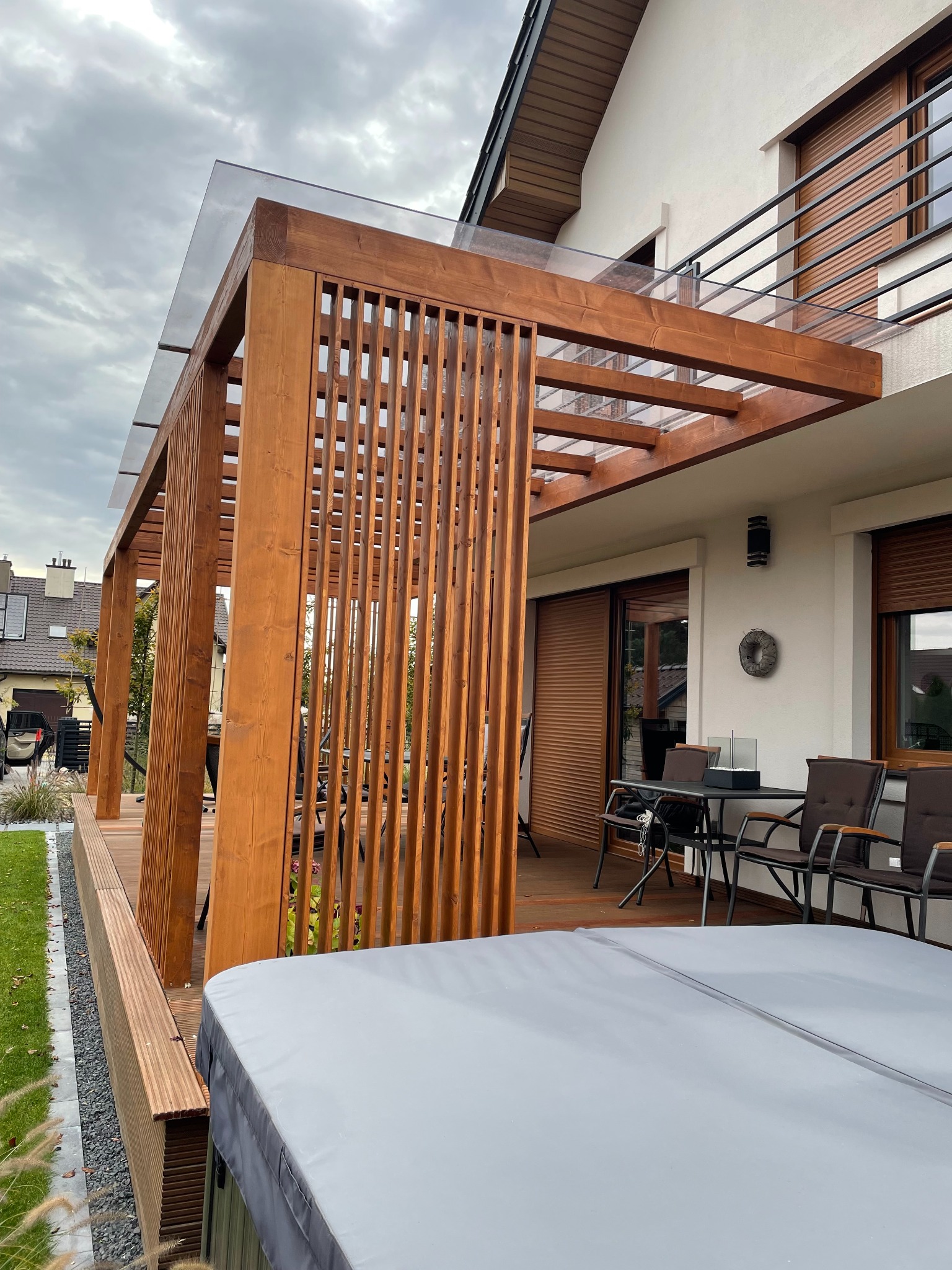 Nowoczesna pergola z drewna i szkła na tarasie, z widocznym jacuzzi i meblami ogrodowymi. Eleganckie połączenie naturalnych materiałów i minimalistycznego designu.