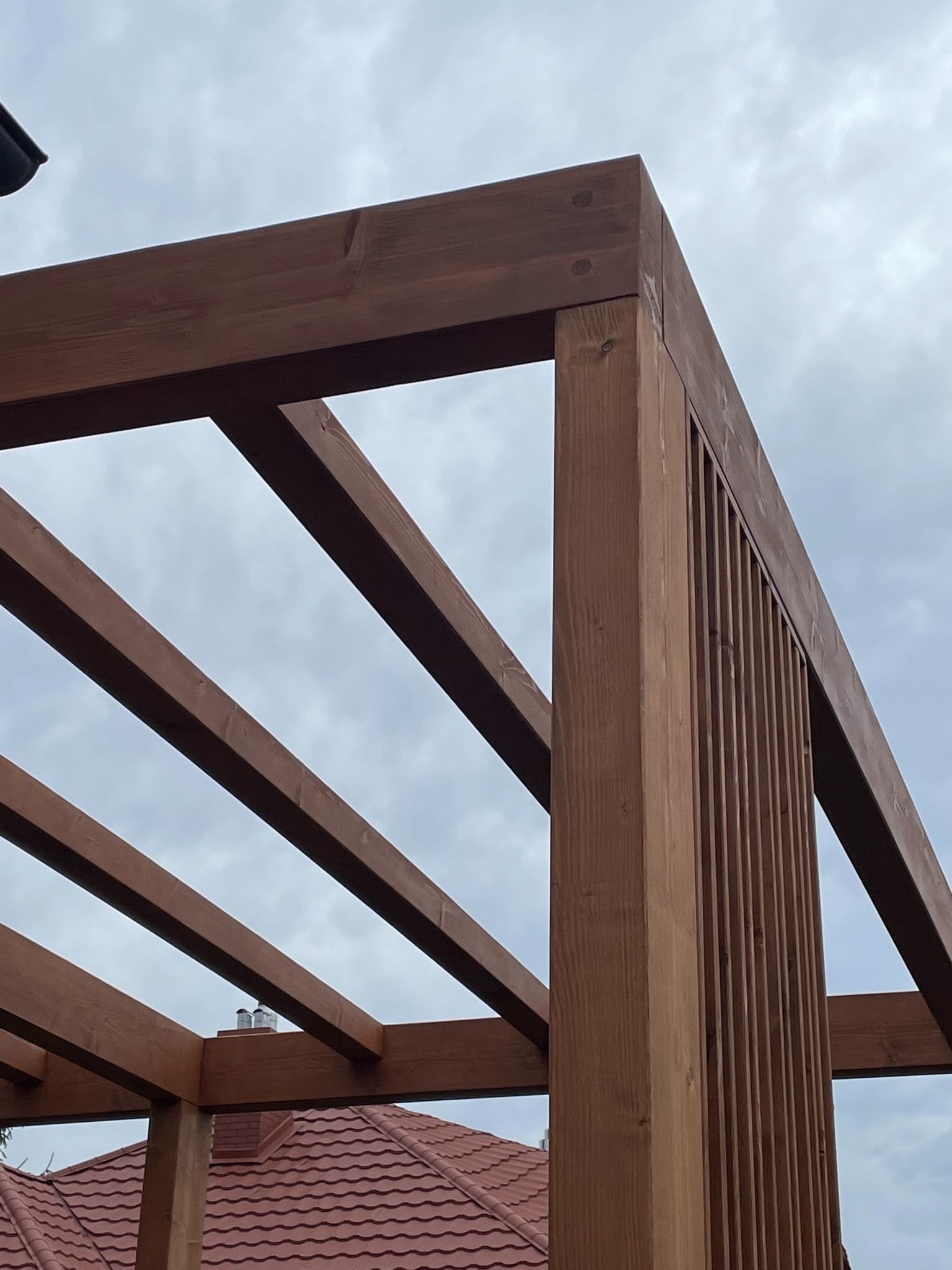 Fragment konstrukcji drewnianej pergoli z widocznymi elementami łączeń i zadaszeniem w tle, wykonana z ciemnego drewna na tle pochmurnego nieba.