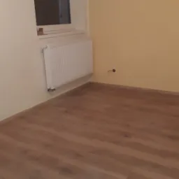 Pusty pokój z panelami podłogowymi w odcieniach brązu, białym grzejnikiem pod oknem i beżowymi ścianami, widoczne gniazdka elektryczne.