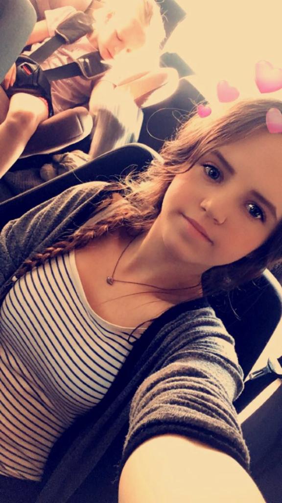 Autoportret młodej kobiety z warkoczem, siedzącej w samochodzie obok śpiącego dziecka w foteliku, z filtrem różowych serduszek na głowie.