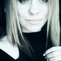 Portret młodej kobiety o jasnych włosach i niebieskich oczach, z piercingiem w nosie, uśmiechającej się delikatnie, na ciemnym tle.