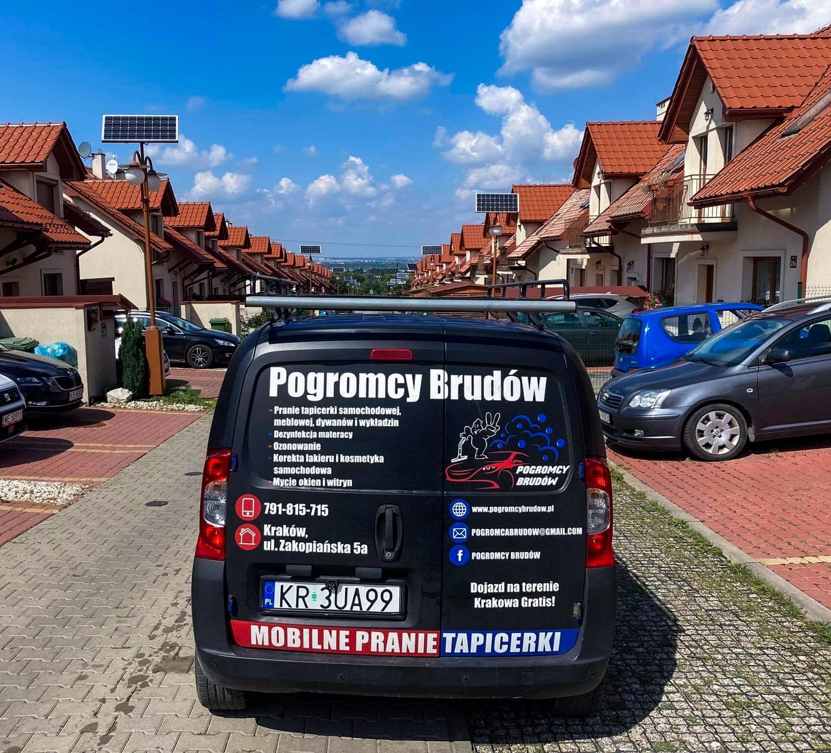 Czarny samochód firmy Pogromcy Brudów z reklamą mobilnego prania tapicerki na tle osiedla domów jednorodzinnych w słoneczny dzień.
