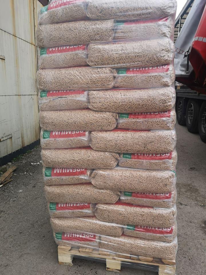 Wysoki stos worków z pelletem drzewnym premium, zapakowanych w przezroczystą folię, ułożonych na palecie drewnianej, z widocznym fragmentem ciężarówki w tle.