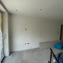 Mebel-Ol - Wnętrze w trakcie remontu: ściany przygotowane do malowania, widoczne puszki elektryczne, podłoga z wylewką, okno zabezpieczone folią malarską, płyta gipsowa oparta o ścianę.