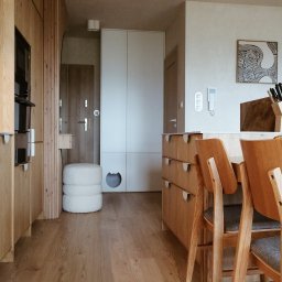 Mebel-Ol - Jasna kuchnia z drewnianymi frontami, minimalistyczna zabudowa, stół z krzesłami i dekoracje. W tle biała szafa z otworem dla kota i wejście do mieszkania.