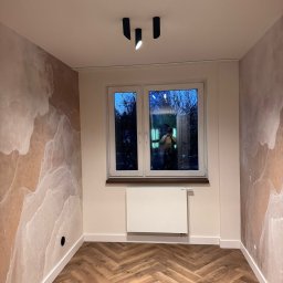 Mebel-Ol - Pokój po remoncie: parkiet w jodełkę, ściany z efektem marmuru, białe okno z widokiem na zewnątrz, nowoczesne oświetlenie sufitowe. Widoczne detale wykończeniowe.