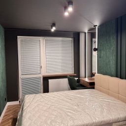 Mebel-Ol - Nowoczesna sypialnia po remoncie: łóżko z beżowym zagłówkiem, biurko z krzesłem, okno z żaluzjami, ciemne ściany z zielonym akcentem, oświetlenie punktowe.