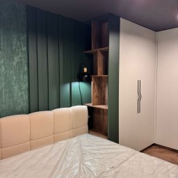 Mebel-Ol - Nowoczesna sypialnia z łóżkiem o beżowym wezgłowiu, zieloną ścianą z lameli, drewnianą półką i białą szafą z czarnymi uchwytami. Całość oświetlona punktowo.
