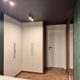 Mebel-Ol - Nowoczesna szafa wnękowa z białymi frontami i minimalistycznymi uchwytami, uzupełniona drewnianymi lamelami. Parkiet w jodełkę i ciemny sufit dodają elegancji.