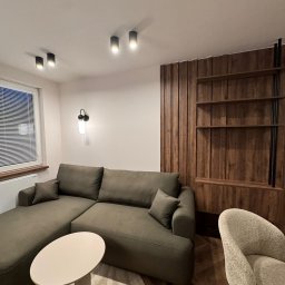 Mebel-Ol - Nowoczesny salon z oliwkową sofą narożną, okrągłym stolikiem i drewnianą zabudową ściany z półkami. Minimalistyczne oświetlenie sufitowe i designerski fotel.