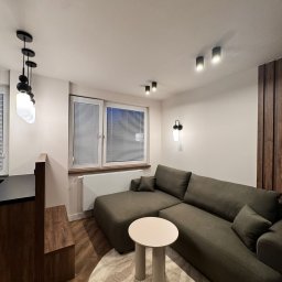 Mebel-Ol - Nowoczesny salon z narożną sofą w odcieniach zieleni, okrągłym stolikiem i drewnianą ścianą. Oświetlenie punktowe i wiszące lampy dodają wnętrzu przytulności.