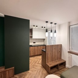 Mebel-Ol - Nowoczesna kuchnia z drewnianymi frontami i białymi szafkami wiszącymi, oddzielona od salonu zieloną ścianką działową i drewnianym barkiem z siedziskiem.