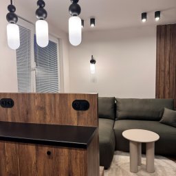 Mebel-Ol - Nowoczesny salon z drewnianą zabudową, szarą sofą i okrągłym stolikiem na wzorzystym dywanie. Oświetlenie stanowią wiszące lampy i reflektory sufitowe.