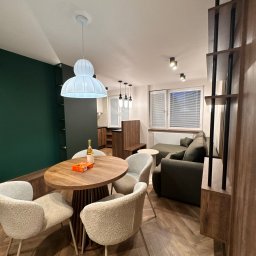 Mebel-Ol - Nowoczesne wnętrze: drewniany stół z krzesłami, zielona ściana, sofa i regał z drewna. Parkiet ułożony w jodełkę. Lampa wisząca nad stołem.