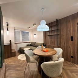 Mebel-Ol - Nowoczesne wnętrze: drewniana podłoga w jodełkę, okrągły stół z krzesłami, sofa w tle, drewniana ściana z półkami i designerskie oświetlenie.