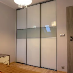 Wysoka szafa przesuwna z białymi i szarymi szklanymi panelami, wbudowana w sypialni z drewnianą podłogą i lampą sufitową z kryształkami.