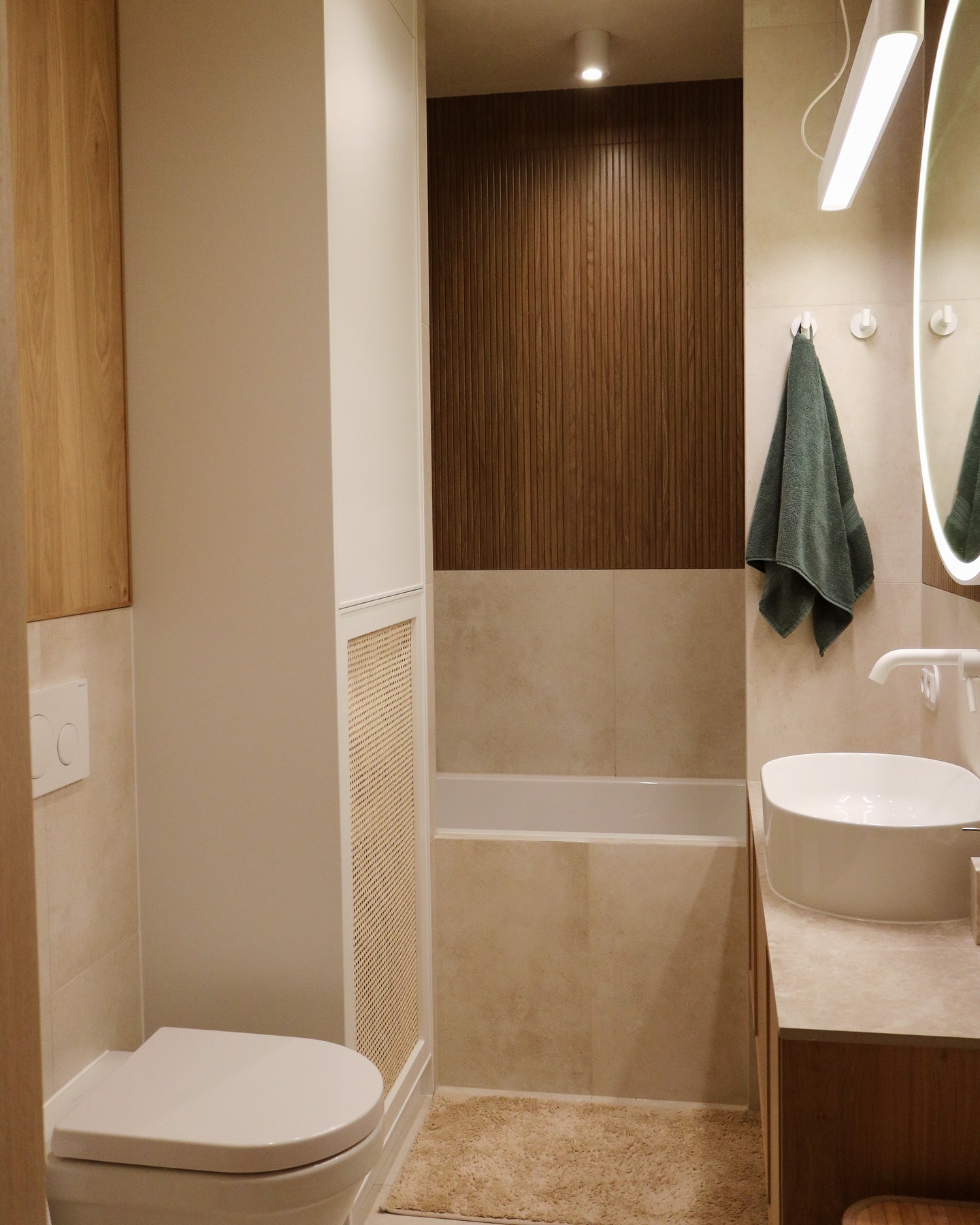 Nowoczesna łazienka z drewnianymi akcentami: panel ścienny, szafka pod umywalką i zabudowa. Umywalka nablatowa, wanna w zabudowie, beżowe płytki i tekstylia.