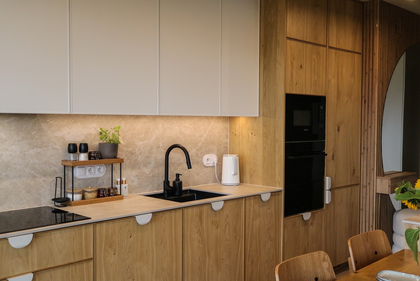 Nowoczesna kuchnia z drewnianymi frontami, czarną armaturą i białymi szafkami górnymi. Uwagę przykuwa marmurowy backsplash i minimalistyczne uchwyty.