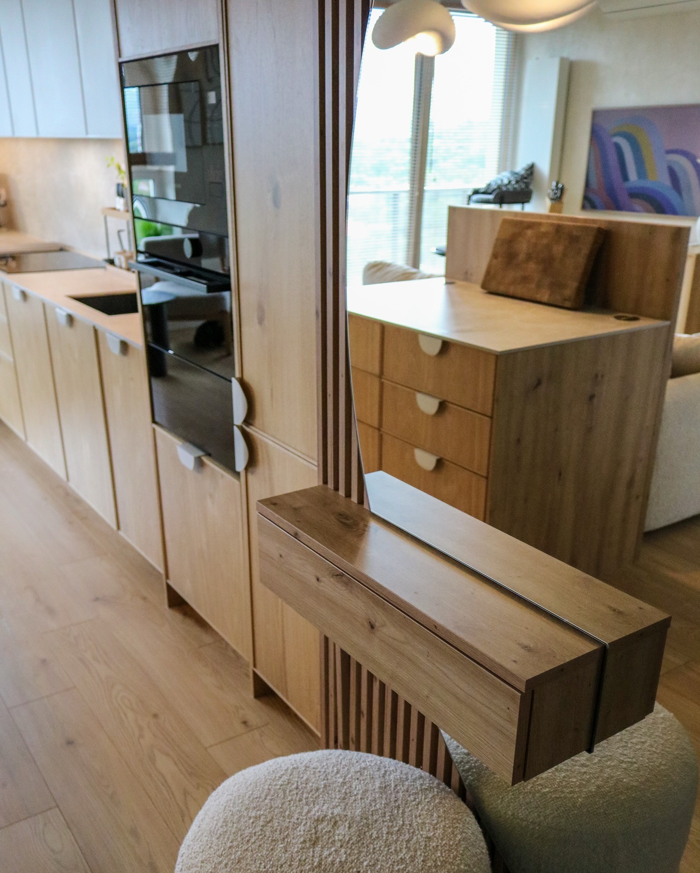 Jasna, drewniana zabudowa kuchenna z piekarnikiem i wysuwaną szafką, odbijająca w lustrze fragment biurka i okna. U dołu pufa z jasnym obiciem.